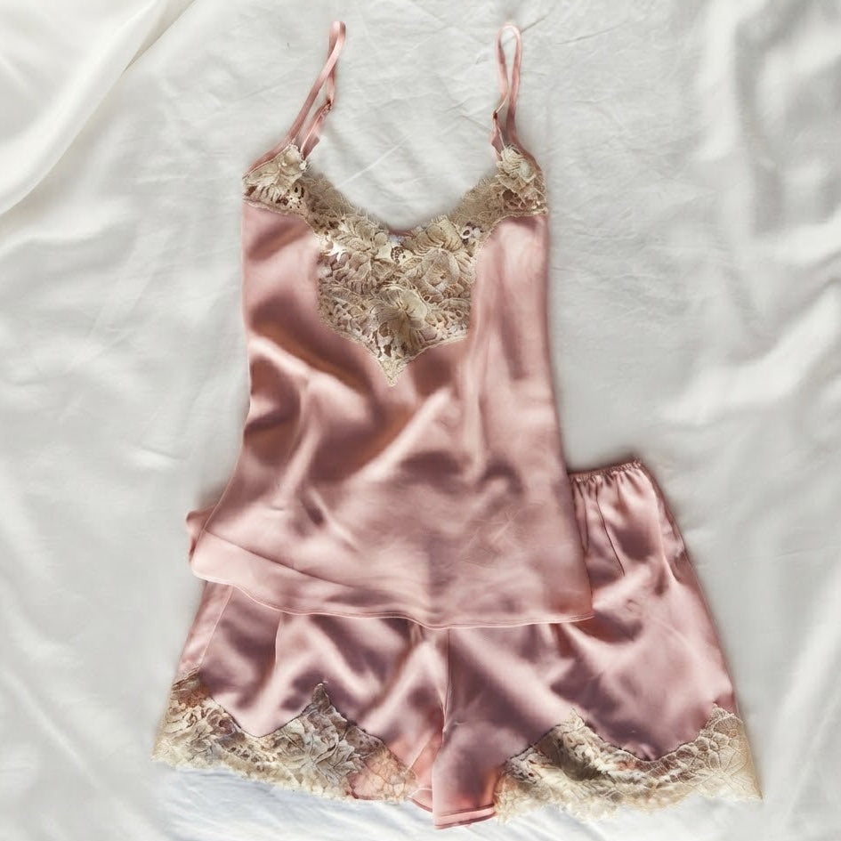 A two-piece satin pajama set in dusty pink, featuring a camisole top and matching shorts with beige floral lace trim, laid flat on a white fabric background.