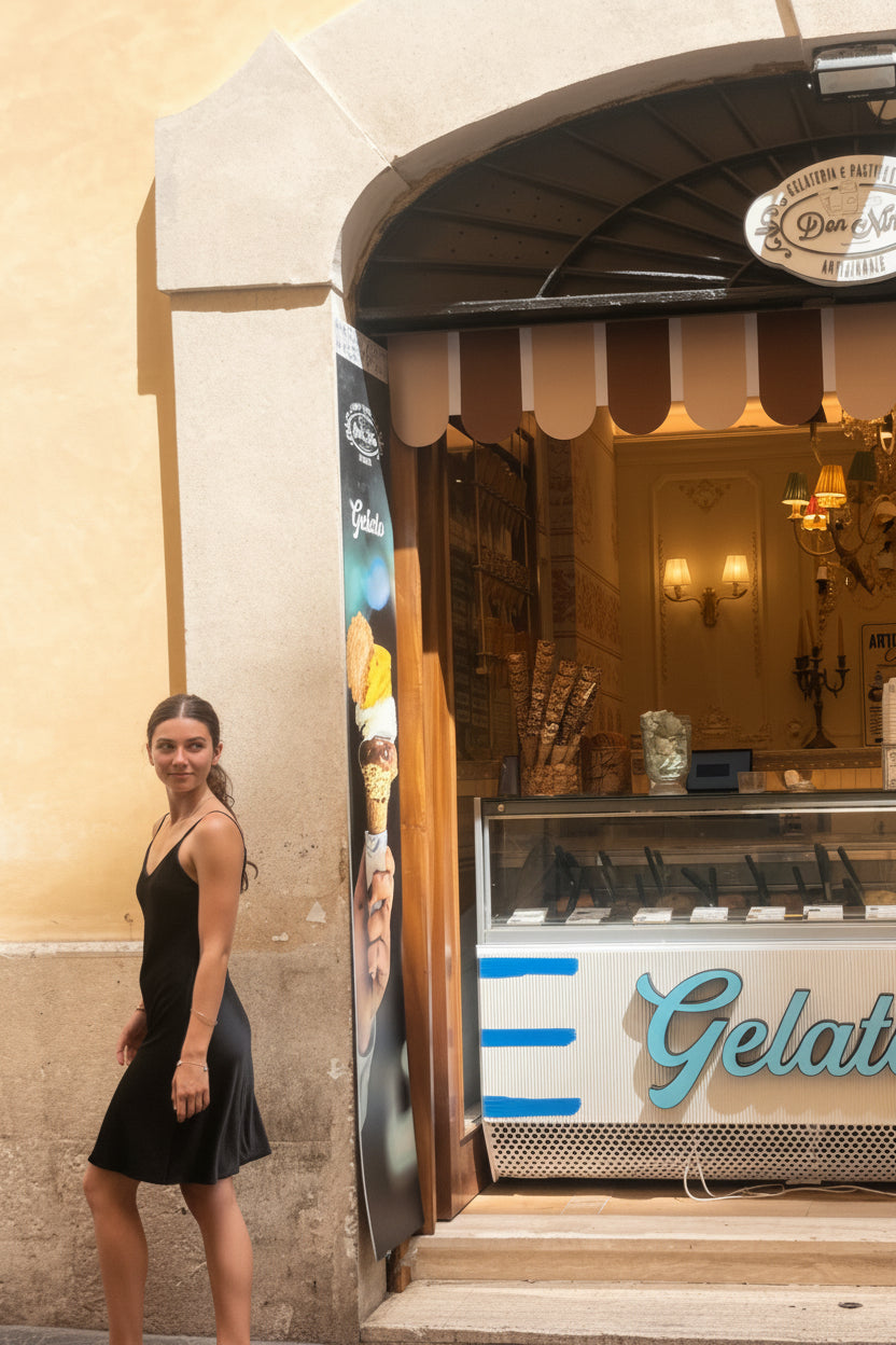 A woman in a black dress walks past a gelato shop storefront on a cobblestone street. The shop has a striped awning, a display case, and a sign that reads 'Gelato'.