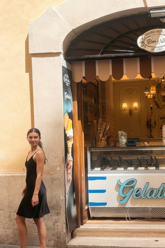 A woman in a black dress walks past a gelato shop storefront on a cobblestone street. The shop has a striped awning, a display case, and a sign that reads 'Gelato'.