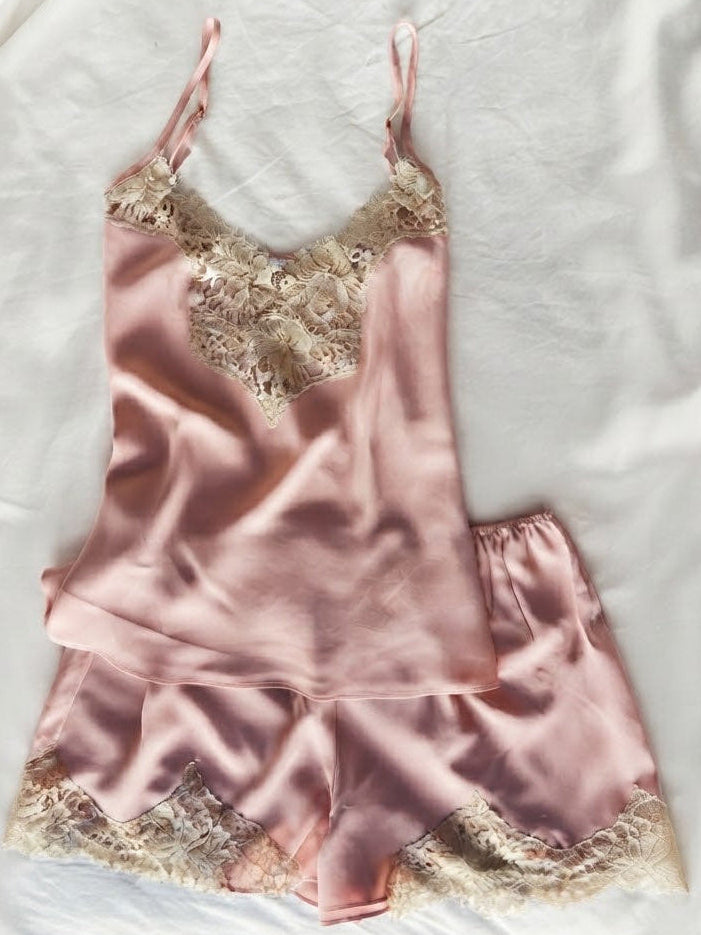 A two-piece satin pajama set in dusty pink, featuring a camisole top and matching shorts with beige floral lace trim, laid flat on a white fabric background.